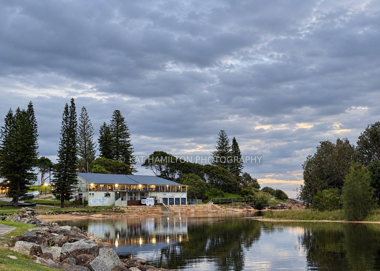 Coastal waterfront buildings and tall pine trees reflected in calm water beneath a cloudy evening sky.