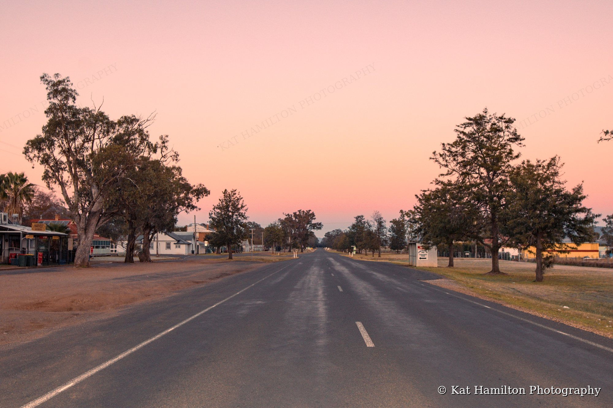 An empty country town street at dawn beneath a pink and peach sky, with gum trees on either side.