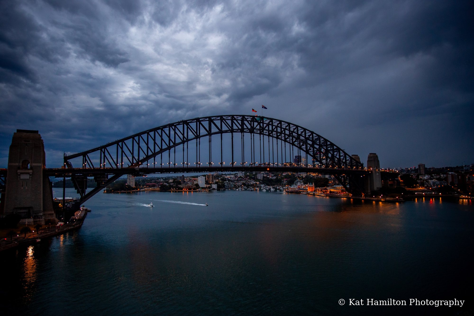 The Sydney Harbour Bridge under heavy storm clouds at dusk, with city lights reflected in the water.