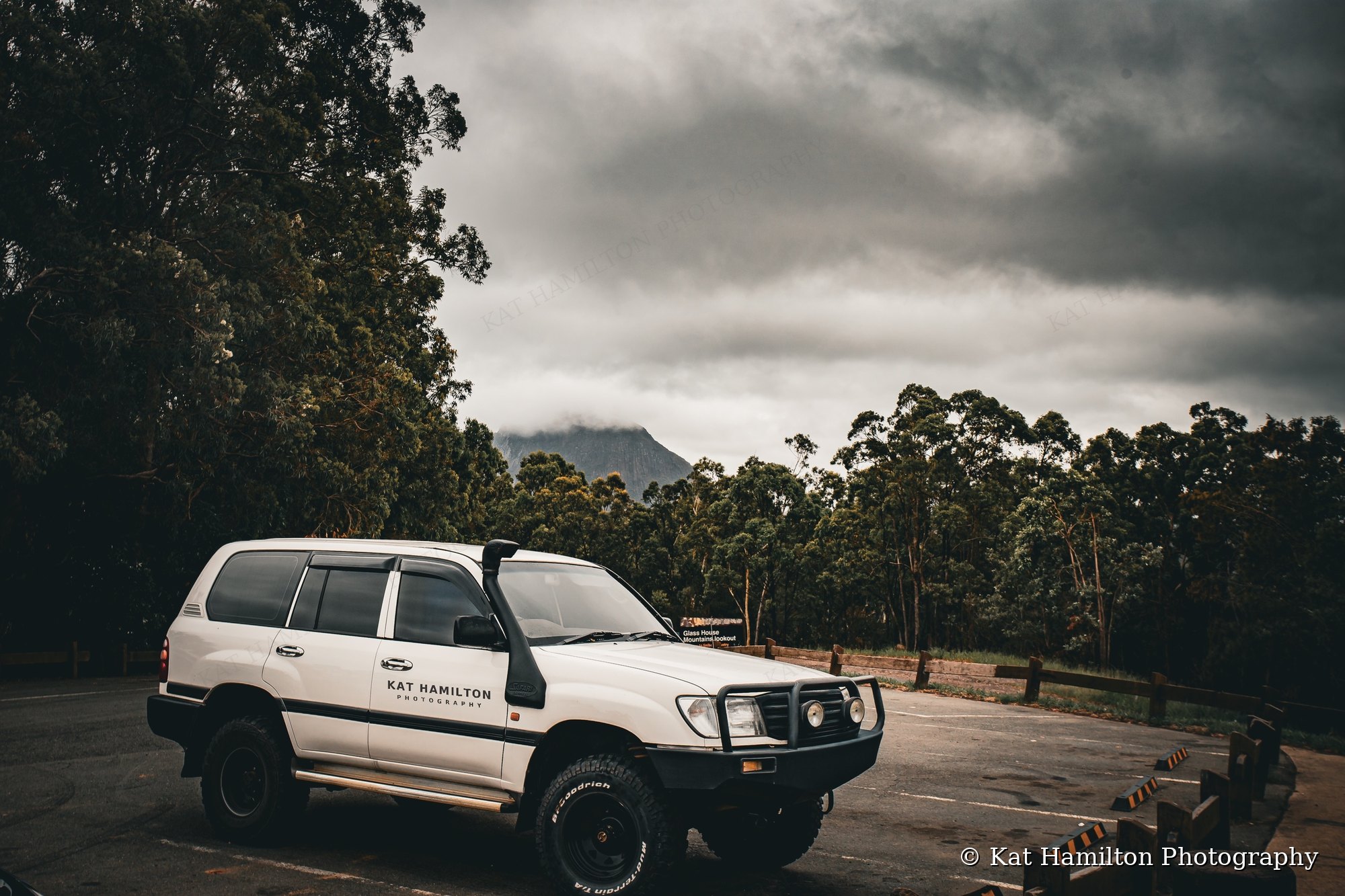 A white LandCruiser with Kat Hamilton Photography lettering on the door, parked at a lookout beneath moody clouds with a mountain peak behind the trees.