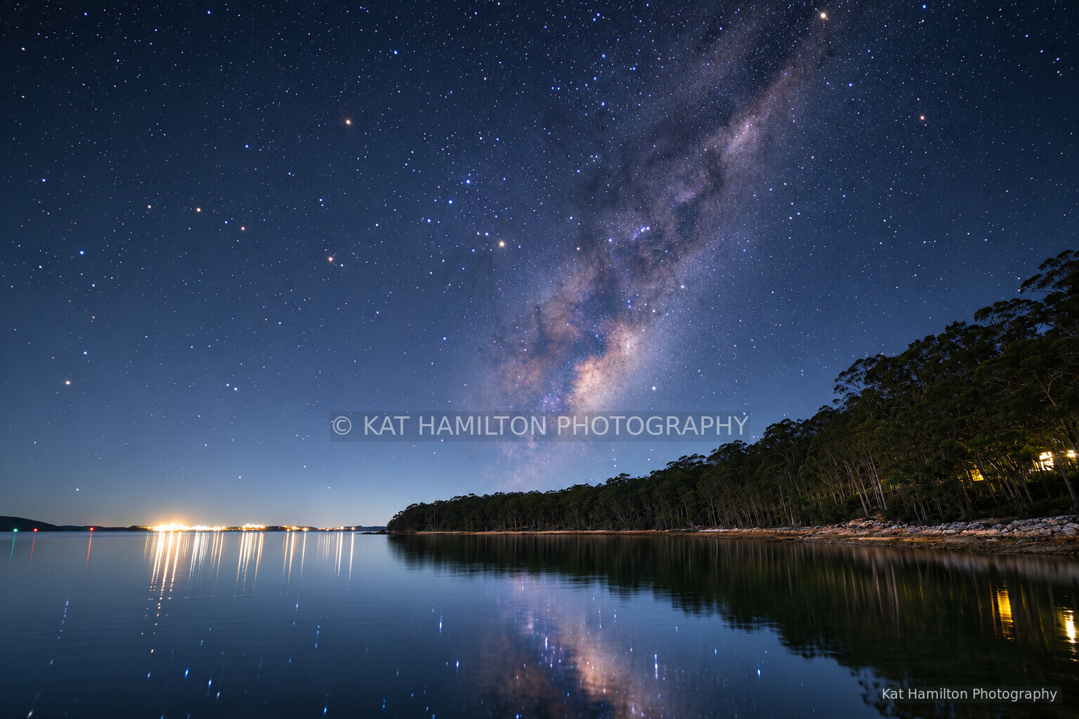 The Milky Way reflected in calm water beside a tree-lined shore, with distant lights along the horizon.